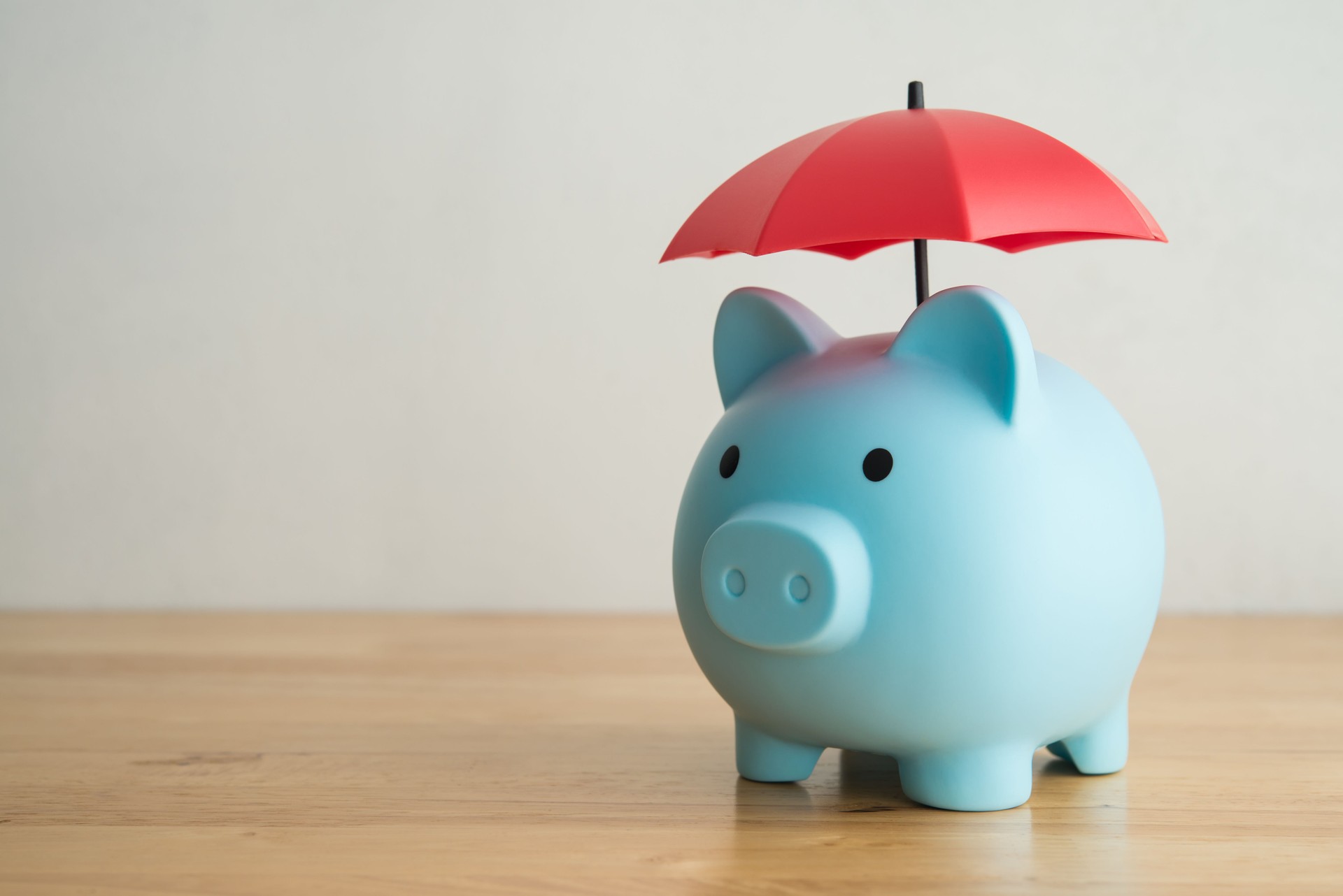 Blue piggy bank under red umbrella on wooden table with white wall background copy space.